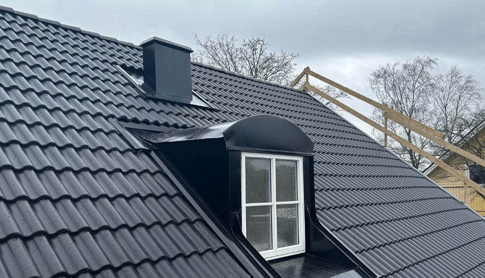 Närbild av ett mörkfärgat plåttak, tillverkat med expertplåtslageri, med skorsten och liten takkupa under en molnig himmel. I bakgrunden syns kala träd och en träkonstruktion.