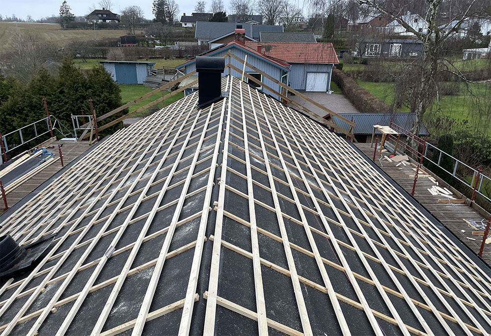Vy från ovan över ett Jono tak-tak under uppbyggnad, som visar ett rutnät av träreglar över svart underlagstak, omgivet av byggnadsställningar, med hus och träd i bakgrunden.